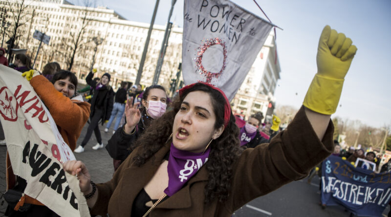 Im Bild: Eine Frau reckt eine Faust in die Höhe, sie trägt Putzhandschuhe. Im Hintergrund ein Schild "Power to the women".