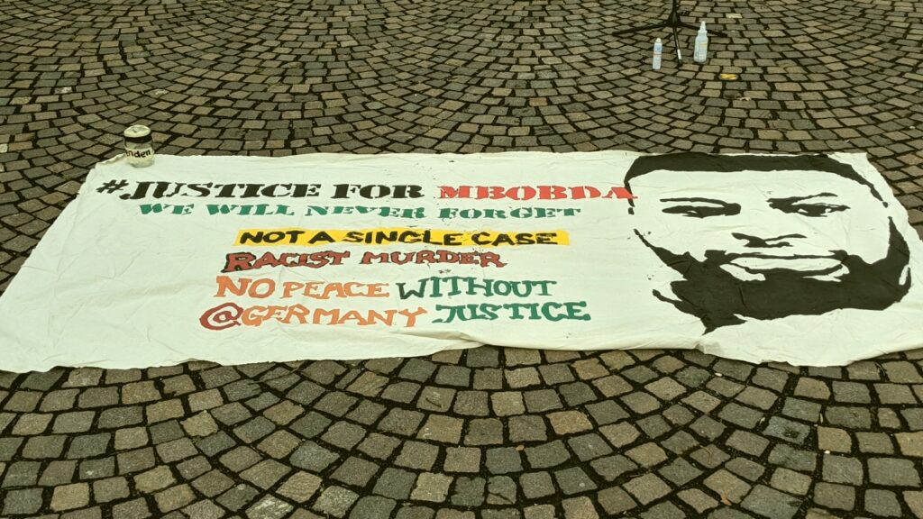 Foto: Kundgebung zum Black History Month am 27.02.21: Transparent mit der Aufschrift "Justice for Mbobda. We will never forget. Not a single case. Racist Murder. No Peace Without @germany justice.