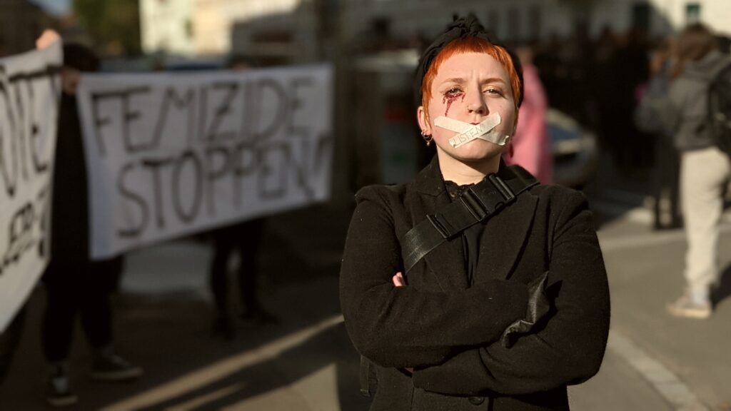 Foto: Eine Person hat sich die Lippen mit Klebeband zugeklebt, auf dem Klebeband steht "Listen" im Hintergrund das Transparent "Femizide stoppen"